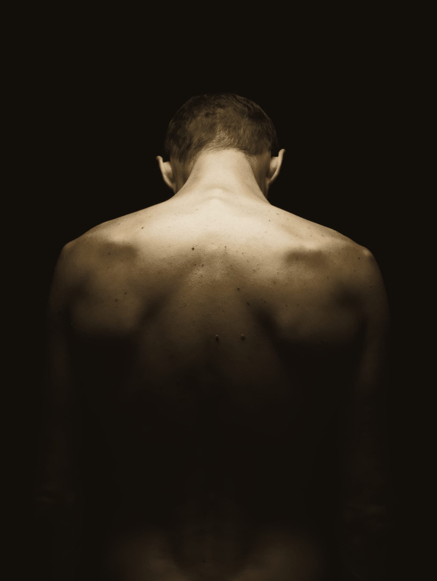 A figure seen from behind, shoulders and back lit by warm side light against a dark ground.