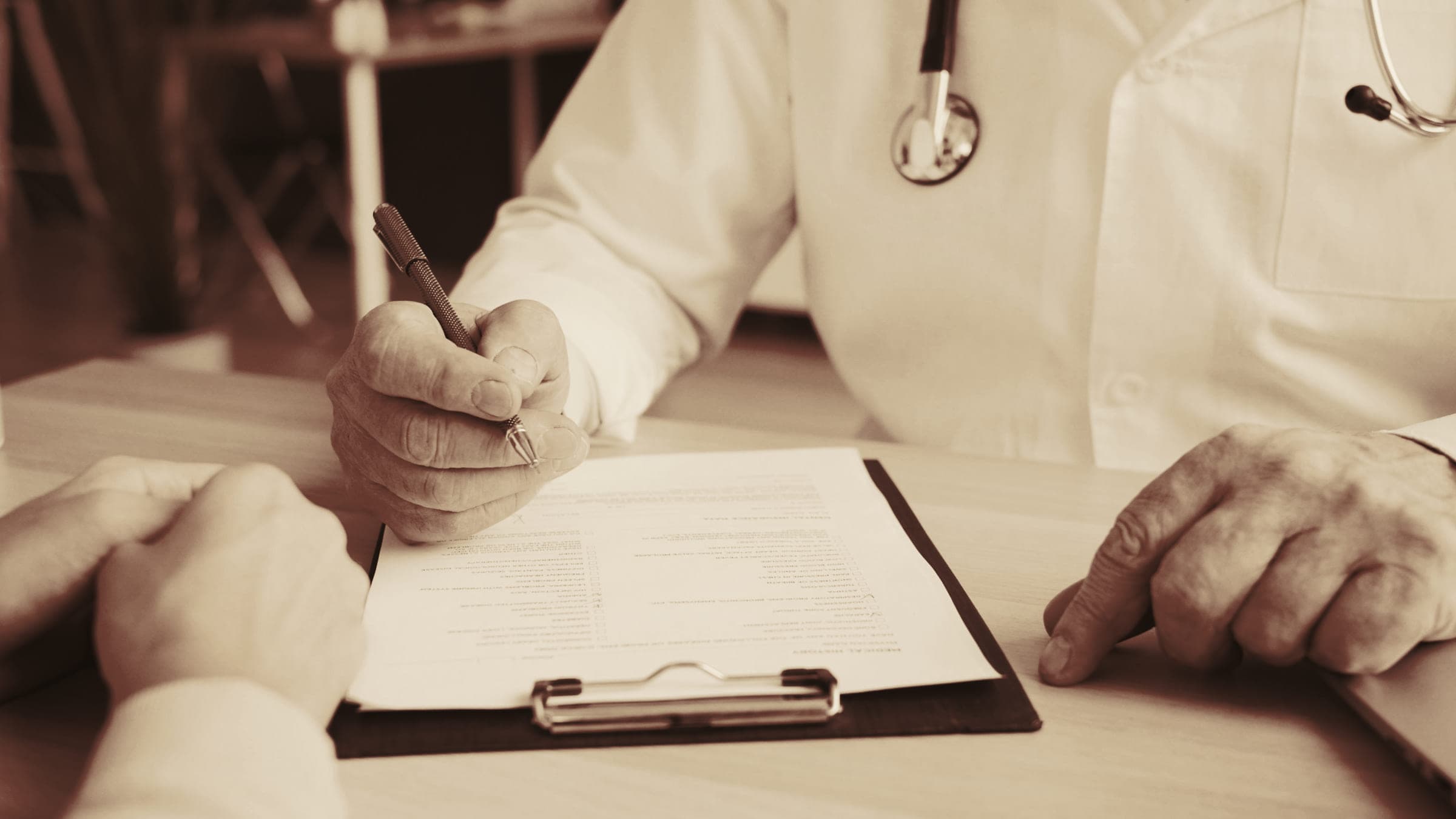 A doctor writing on a clipboard across a desk from a patient; neither face is in frame.